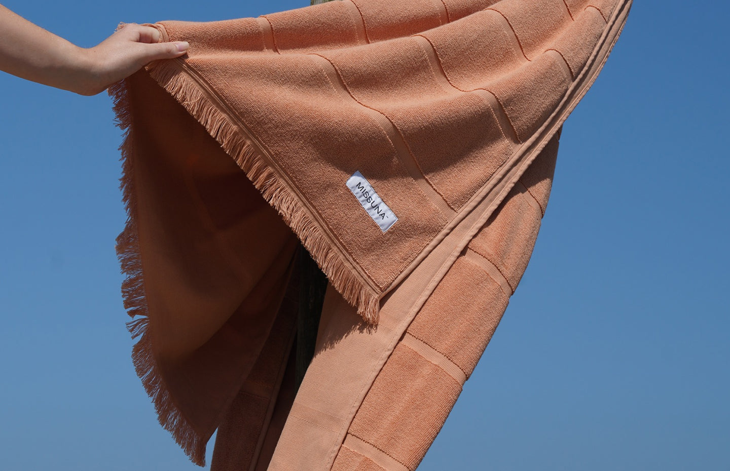 Hand holding a textured cotton towel with fringed edges against a clear blue sky.